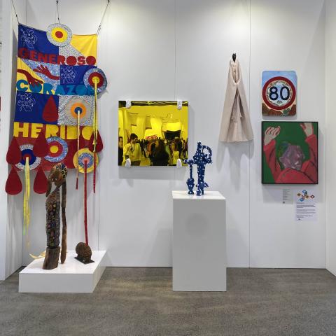 5 contemporary art pieces hang on a wall of a temporary gallery space. Two pieces are on plinths. On the wall there is hanging textile in primary colours, a square yellow painting showing people in a crowded room, a small  beige fabric piece, a square pai