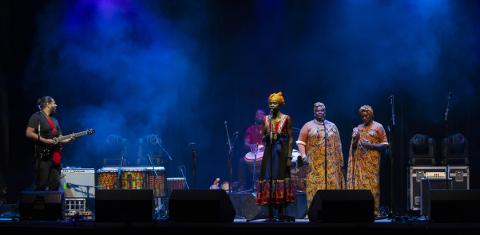 This image shows three singers in colourful, patterned clothing, with a guitarist and drummer, on a lit performance stage as part of the opening night of the 2021 Castlemaine State Festival. Ajak Kwai and Riel Dë Nyiër: World premiere at the Castlemaine 