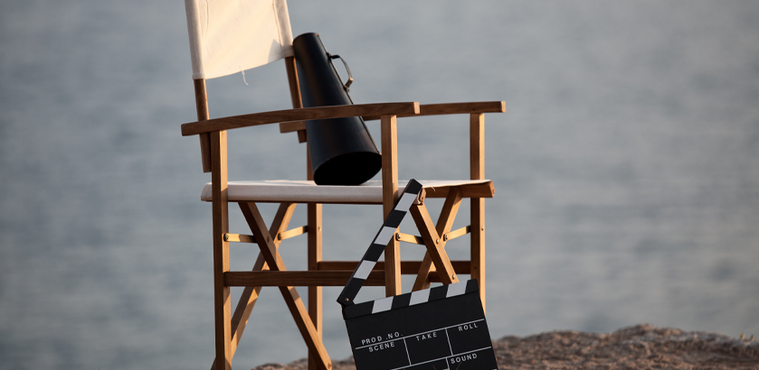 Director's chair with a megaphone sitting on it and a film board standing on the ground nearby. There is a water scene in the background. Could be a lake or sea.