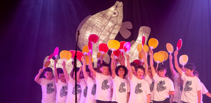 Primary school aged boys sing in a choir. They wear white t-shirts with black patterns and hold up paddles. A white fish sculpture is lit-up behind them. 