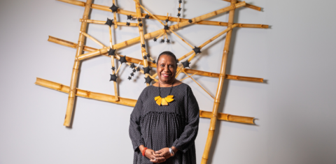 Artist, Gail Mabo, stands in front of artwork in a gallery. Artwork is made from timber and is adorned with black stars.