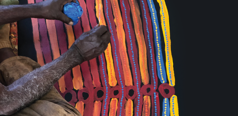 An artist's hands, holding a paint jar and painting tool, working on a colourful Indigenous artwork, which features yellow, blue, orange and red designs. 