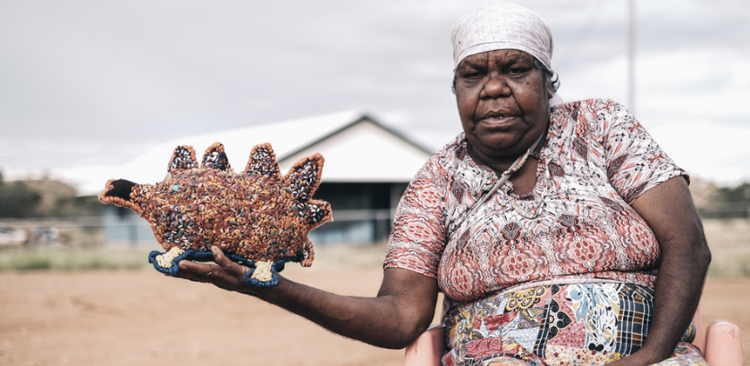 First Nations artist Marlene Rubuntja wears a dress with red, white, balck and yellow designs. She sits in a rural setting with a shed in the background, she is looking at the camera with one arm outstretched hold an echidna she stitched.