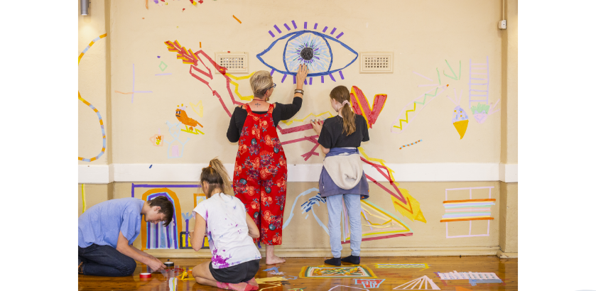 A woman and three teenagers creating art on the wall and floor using colourful tape.