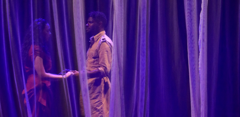 First Nations male actor holds hands with a female actor on stage during a performance. Sheer curtains surround them and the stage is lit in purple lights.