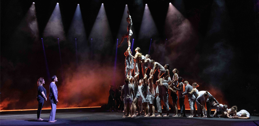 Theatre performers in a human pyramid with two people looking on from a distance.