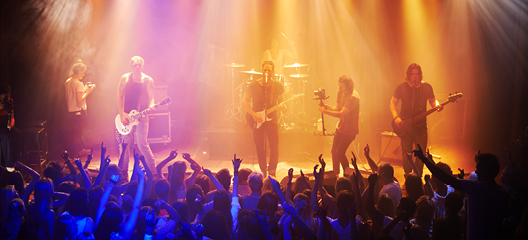  A rock band performs live on stage, illuminated by vibrant, colourful stage lights. A cheering crowd, with hands raised, enjoys the energetic performance.