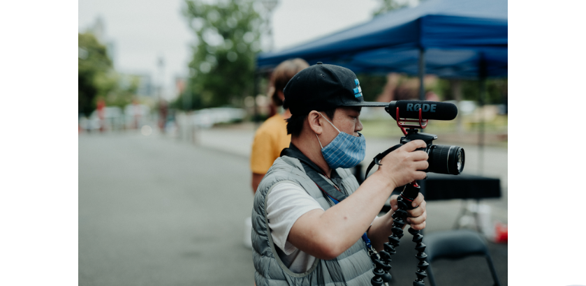 Vinh Nguyen wears a baseball cap and facemask and holds a video camera with microphone and tripod.