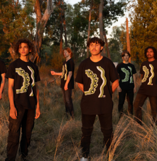 Group of First Nations choir boys are pictured standing in a bush landscape at dusk. They wear black and green uniforms. 