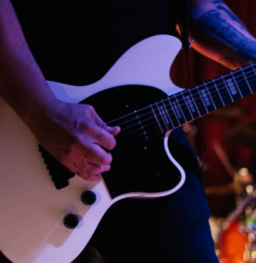 A close up of a person playing a white electric guitar on stage with an amp, to the left, and a drummer to the right.